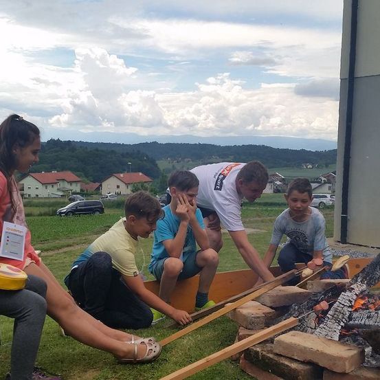 Eine Gruppe von Kindern und Erwachsenen versammelt sich um ein Feuer im Freien. Sie bereiten sich darauf vor, Essen am Feuer zu kochen oder zu rösten.