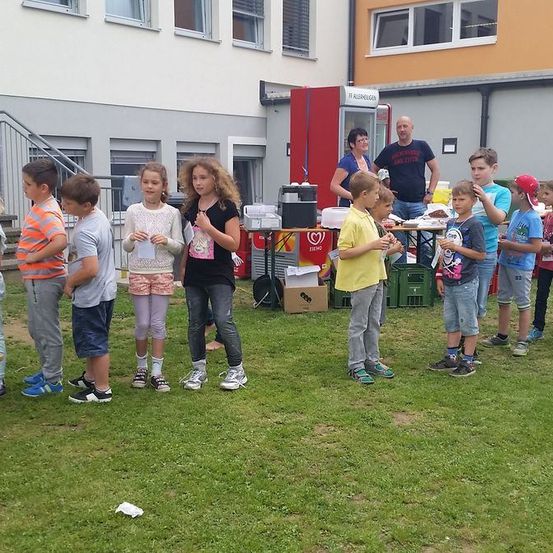 Eine Gruppe von Kindern steht auf dem Rasen vor einem Gebäude. Ein Mann und eine Frau stehen hinter einem Tisch mit Essen.