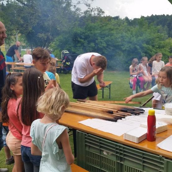 Eine Gruppe von Kindern und Erwachsenen steht um einen Tisch mit Holzstäben. Ein Junge bläst auf die Stäbe, während andere zusehen. In der Nähe gibt es Bänke und einen Kinderwagen. Im Hintergrund sind Bäume und ein Gebäude zu sehen.