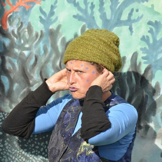 Eine Frau mit blauer Farbe im Gesicht, die eine grüne Mütze trägt, steht vor einem Hintergrund mit gemalten Korallenriffen.