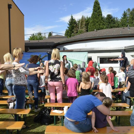 Eine Gruppe von Menschen, darunter Erwachsene und Kinder, versammelt sich im Freien. Sie sitzen auf Bänken, und einige stehen. Eine Person führt eine Zaubershow vor ihnen auf. Der Bereich ist von Bäumen umgeben, und im Hintergrund befindet sich ein Gebäude.