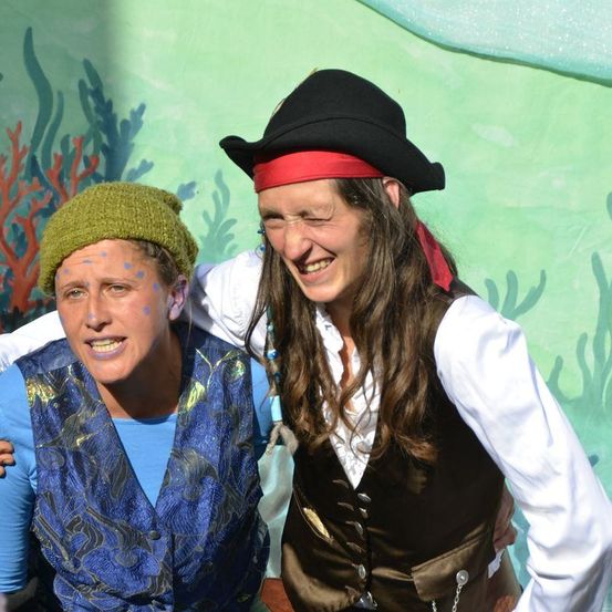 Zwei Frauen, die als Piraten gekleidet sind, stehen vor einer grünen Wand mit Korallenriff-Designs. Die Frau auf der linken Seite trägt ein blaues Shirt und einen grünen Hut, während die Frau auf der rechten Seite einen schwarzen Hut und ein rotes Stirnband trägt. Sie lächeln beide.