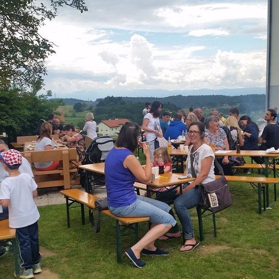 Menschen versammeln sich in einem Außenbereich, um eine Mahlzeit zu genießen. Einige sitzen an Tischen, während andere stehen und reden. Die Umgebung besteht aus einer Rasenfläche, Bänken und Tischen. Der Hintergrund zeigt Bäume, Gebäude und eine malerische Aussicht.