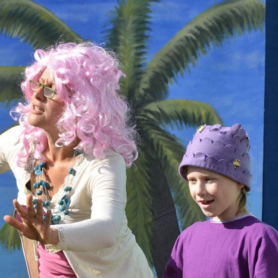 Eine Frau mit pinker Lockenperücke und ein junges Mädchen mit einem lila Hut stehen vor einer gemalten Palme.