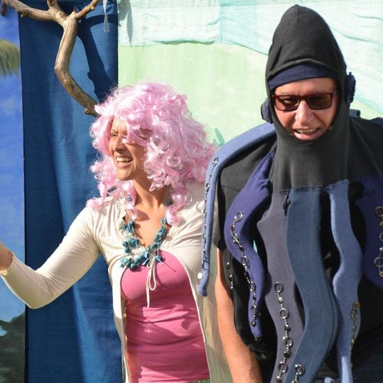 Eine Frau mit rosa lockigem Haar und einem rosa Oberteil steht neben einem Mann in einem blauen und schwarzen Kostüm. Sie lächeln beide und scheinen für ein Foto zu posieren.