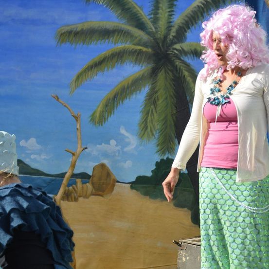 Eine Frau mit rosa Locken steht auf einer Bühne, gekleidet in ein pinkes Oberteil, grüne Hosen und eine weiße Strickjacke. Hinter ihr befindet sich eine gemalte Kulisse einer Strandszene mit einer Palme und einem Stock. Eine weitere Person sitzt auf der linken Seite.