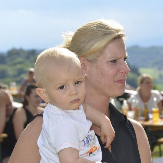 Eine Frau hält ein Baby im Freien in einem Park und sieht mit ernster Miene nach vorne. Hinter ihnen stehen Tische mit Flaschen und Essen, und es sitzen Menschen. Ein ferner Blick auf Bäume und Berge.