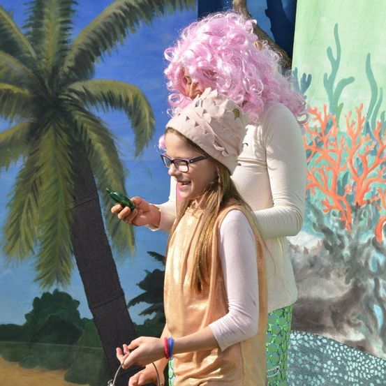 Ein junges Mädchen in einem Kostüm steht neben einer Frau mit rosa Perücke und hält ein grünes Objekt, vor einem Hintergrund aus gemalter Palme und Korallenriff.