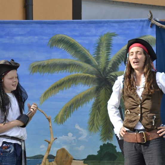 Zwei Menschen in Piratenkostümen stehen vor einem gemalten Hintergrund. Einer hält einen Stock und lächelt, während der andere mit ernster Miene in die Kamera schaut.