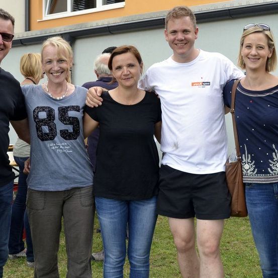 Fünf Menschen stehen in einem Grasbereich vor einem Gebäude. Sie alle haben glückliche Gesichtsausdrücke. Der Mann auf der linken Seite trägt eine Sonnenbrille und ein schwarzes T-Shirt. Die Frau neben ihm trägt ein graues T-Shirt mit der Nummer 85.