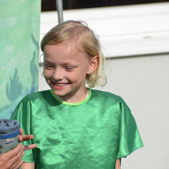 Ein lächelndes Mädchen mit blondem Haar trägt ein grünes Kleid und eine Perlenkette. Eine Person, die einen Becher mit einem Design hält, steht neben ihr.