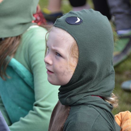 Ein Kind mit einer grünen Kapuze schaut zur Seite. Hinter ihm sitzt eine Person in Grün auf dem Gras.