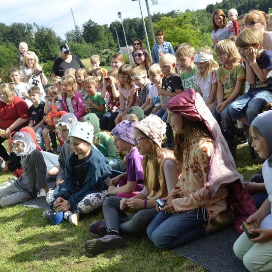 Eine Gruppe von Kindern in Kostümen, einige mit Masken, sitzen auf dem Gras in einem Außenbereich. Einige von ihnen halten etwas in den Händen.