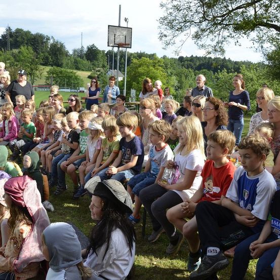 Eine Gruppe von Kindern und Erwachsenen sitzt auf dem Rasen. Einige tragen Hüte, Brillen und Turnschuhe. Das Gebiet ist von Bäumen und einem Basketballkorb umgeben. In der Ferne befindet sich ein Grasfeld mit einem Gebäude und einem Turm.