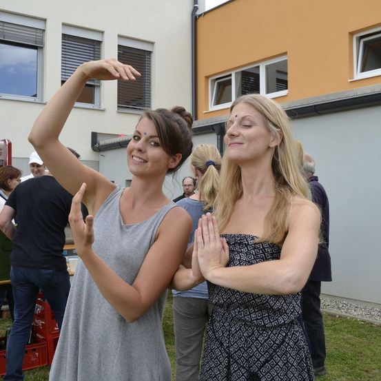 Zwei Frauen stehen draußen und praktizieren Yoga. Eine Frau macht eine Yoga-Pose mit erhobenen Händen. Sie stehen vor einem Gebäude mit Glasfenstern. Hinter ihnen stehen Leute.