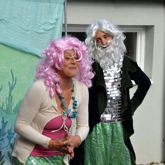 Zwei Menschen in Kostümen, einer mit rosa Haaren und einer Halskette, der andere in einem Paillettenhemd, stehen vor einer weißen Wand.