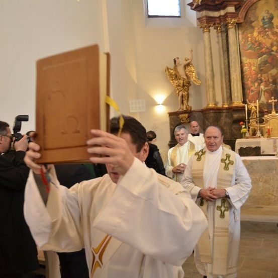 Ein Priester hält ein Buch, während ein anderer hinter ihm in einer Kirche steht, mit einer Statue und einem Altar im Hintergrund.