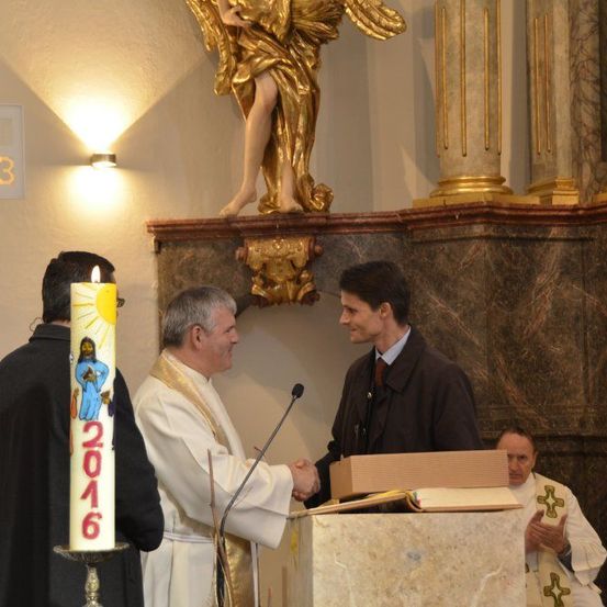 Zwei Männer schütteln sich die Hände vor einem Podium, einer trägt eine weiße Robe und der andere einen dunklen Mantel. Eine goldene Engelsstatue steht hinter ihnen, und eine Kerze mit der Nummer 2016 ist in der Nähe.
