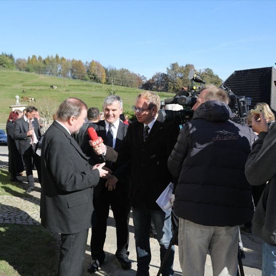 Mehrere Männer in formeller Kleidung sind im Freien, einer gibt ein Interview mit einem Mikrofon. Hinter ihm hält ein Mann eine Kamera und eine Frau filmt. Im Hintergrund steht ein Gebäude und ein Hügel mit Bäumen.