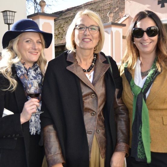 Drei Frauen stehen lächelnd draußen. Die linke Frau hält ein Weinglas. Die Frau in der Mitte trägt eine Brille und eine Lederjacke. Die rechte Frau trägt eine Sonnenbrille und einen grünen Schal.