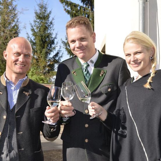 Drei Personen lächeln und halten Weinglas mit Weißwein vor einem Gebäude mit Bäumen im Hintergrund.
