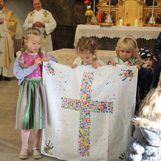 Eine Gruppe junger Mädchen hält ein farbenfrohes Kreuzbanner in einer Kirche, mit Priestern im Hintergrund.