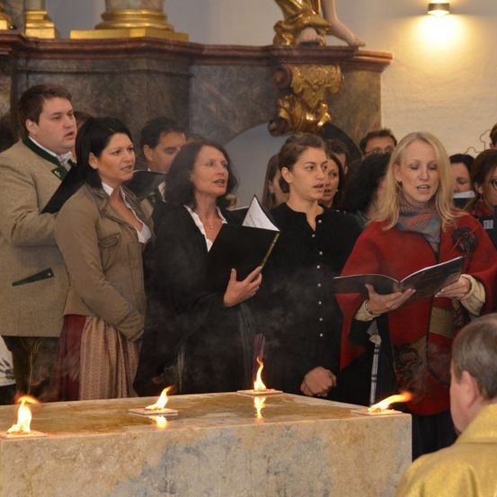 Eine Gruppe von Menschen steht um einen Steinaltar mit brennenden Kerzen in einer Kirche. Einige halten Bücher, während andere zu singen scheinen.
