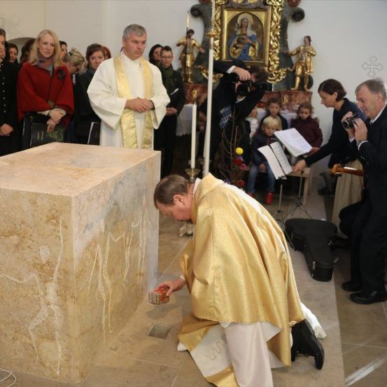 Ein Priester kniet vor einer großen Steinplatte während einer Kirchenzeremonie, während andere die Szene beobachten und fotografieren. Ein großer Rahmen schmückt die Wand hinter dem Altar.