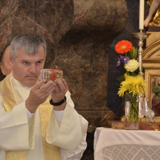 Ein Priester hält ein kleines, kunstvolles Objekt in seinen Händen in einer Kirche. Dahinter steht ein Kerzenständer mit Blumen. Ein Bild einer Frau ist in einem Rahmen in der Nähe.