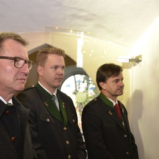 Drei Männer in Uniformen stehen in einem Flur, wahrscheinlich für eine offizielle Veranstaltung. Sie stehen in der Nähe einer Glasscheibe, mit einer Lampe an der Wand befestigt.