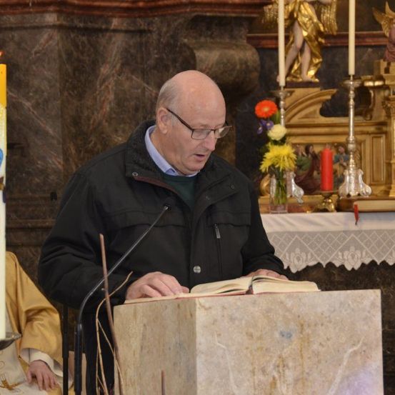 Ein älterer Mann steht in einer Kirche an einem Pult und liest aus einem Buch, mit einer Kerze und Blumen im Hintergrund.