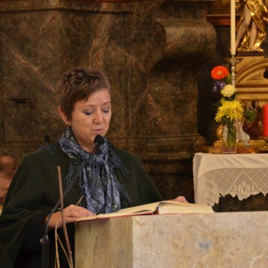 Eine Frau steht an einem Podium, liest ein Buch in ein Mikrofon, mit einem Blumenarrangement und einer Kerze hinter ihr.