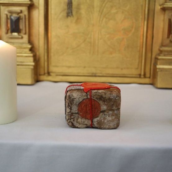 Zwei brennende Kerzen und ein mit Wachs versiegelter Gegenstand stehen auf einem weißen Tuch vor einem goldenen Altar.