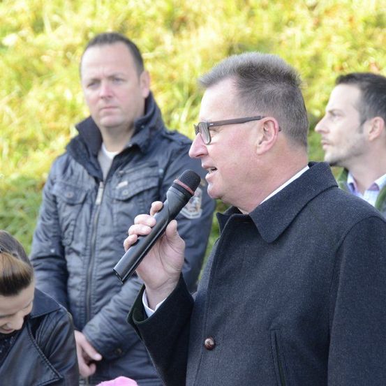 Ein Mann mit Brille spricht in ein Mikrofon. Hinter ihm stehen drei Personen, darunter eine Frau mit einem Pferdeschwanz.