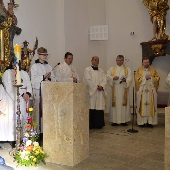 Geistliche stehen in einer Kirche, einige halten Bücher. Ein Podest und ein Mikrofon stehen davor. Eine goldene Kerze steht auf einem Ständer und eine Blumenarrangement ist in der Nähe.