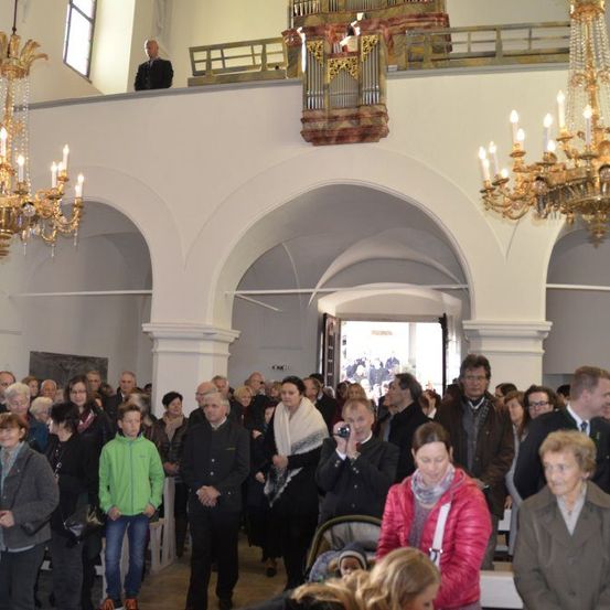 Eine Gruppe von Menschen versammelt sich in einer Kirche, stehend und sitzend. Einige schauen zur Kamera. Kronleuchter hängen von der Decke. Ein Mann steht auf einem Balkon oben, in der Nähe einer Orgel.