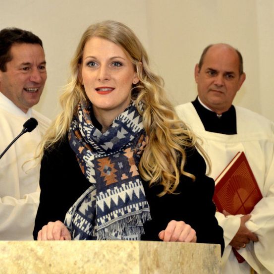 Eine Frau steht an einem Podest und trägt ein gemustertes Tuch. Im Hintergrund stehen zwei Priester, die ein Buch halten und aufmerksam erscheinen. Die Frau schaut direkt in die Kamera.