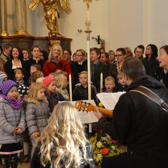 Ein Kinderchor steht in einer Kirche und singt, während ein Mann Gitarre spielt. Kerzen und Blumen stehen vor ihnen, mit einer Engelsstatue dahinter.