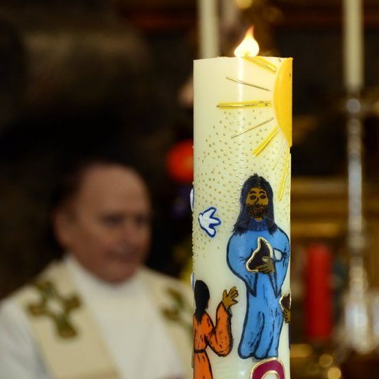 Eine Kerze mit einem gelben Sonnendesign und blauen Illustrationen eines Mannes und eines Kindes brennt. Ein Priester steht dahinter und trägt ein weißes Gewand mit einem grünen Kreuz.