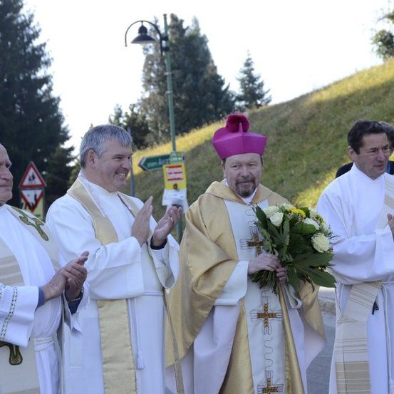 Vier Priester stehen in einer Reihe, einer hält Blumen, in weißen und goldenen Gewändern. Sie lächeln und scheinen zu klatschen. Ein Schild ist hinter ihnen sichtbar, und es gibt Bäume und einen Hügel im Hintergrund.
