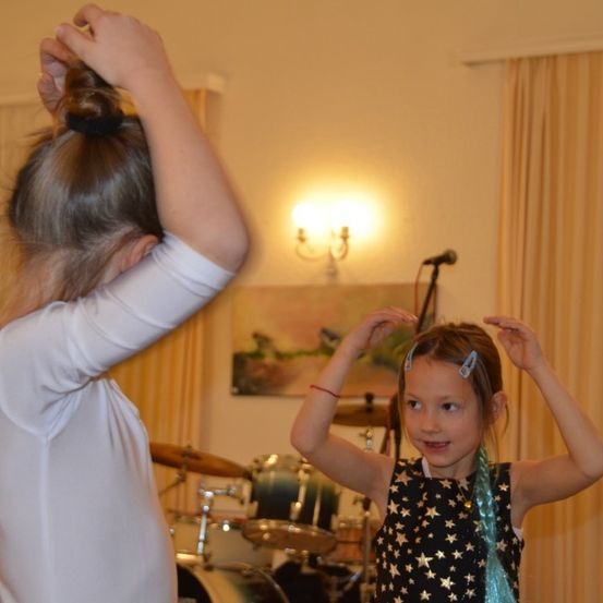 Zwei junge Mädchen tanzen in einem Raum mit Musikinstrumenten. Das Mädchen rechts trägt ein sternenförmiges Kleid.