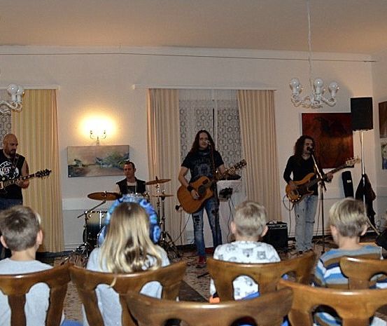 Eine Band spielt in einem Raum mit Schlagzeug, Lautsprechern und Gitarren, während mehrere Kinder von Holzstühlen aus zusehen.
