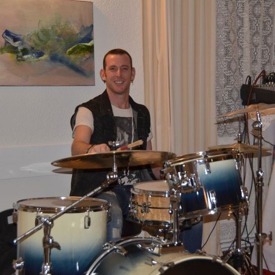 Ein Mann spielt Schlagzeug in einem Raum mit einem Gemälde an der Wand und einem Mikrofon auf einem Ständer. Er lächelt und hält Schlagzeugstöcke in der Hand.
