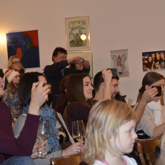 Eine Gruppe von Menschen sitzt in einem Raum mit Weingläsern, einige machen Fotos mit ihren Handys. Der Raum hat Kunst an der Wand, darunter ein gerahmtes Gemälde und ein großes Poster.