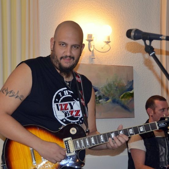 Ein Mann spielt Gitarre in einem Raum mit Mikrofon, einem weiteren Musiker dahinter und einer an der Wand montierten Lampe.