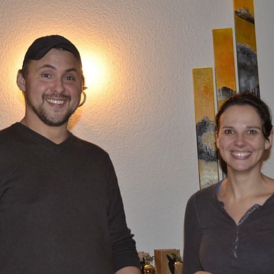 Ein Mann und eine Frau stehen nebeneinander und lächeln. Sie tragen dunkle Oberteile. Hinter ihnen befindet sich eine Wand mit drei farbenfrohen abstrakten Gemälden.