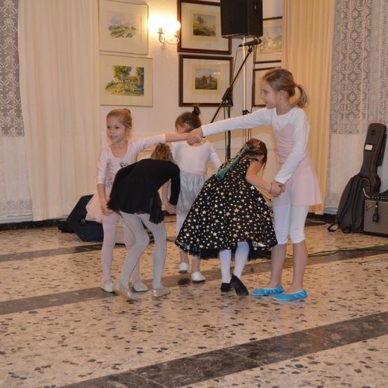 Mehrere junge Mädchen tanzen auf einem gefliesten Boden. Ein Mädchen in Pink hält ein anderes in einem schwarzen und sterngemusterten Kleid. Im Hintergrund sind Musikgeräte zu sehen.