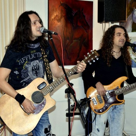 Zwei Männer spielen Gitarren in einem Raum. Einer spielt eine akustische Gitarre und singt in ein Mikrofon, während der andere eine E-Gitarre spielt. Hinter ihnen hängen Pferdegemälde.