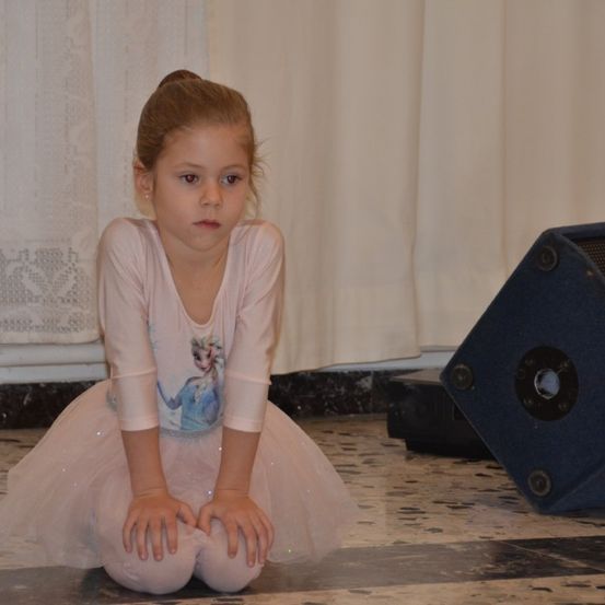 Ein kleines Mädchen in einem rosa Tutu mit einer Frozen-Figur auf dem Shirt kniet auf einem gefliesten Boden und schaut nach vorne, mit einem Lautsprecher im Hintergrund.