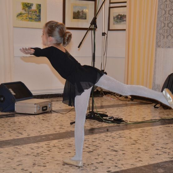 Ein junges Mädchen in einem schwarzen Kleid und weißen Leggings führt eine Ballettpose in einem Raum aus. Sie steht auf einem gefliesten Boden, in der Nähe eines Lautsprechers und von Ausrüstung.